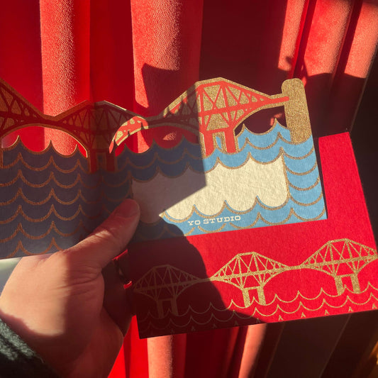Hand holding a decorative card with bridge design against a red curtain background