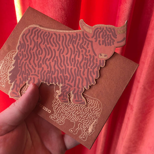 Hand holding a card with a Highland Cow design against a red curtain background