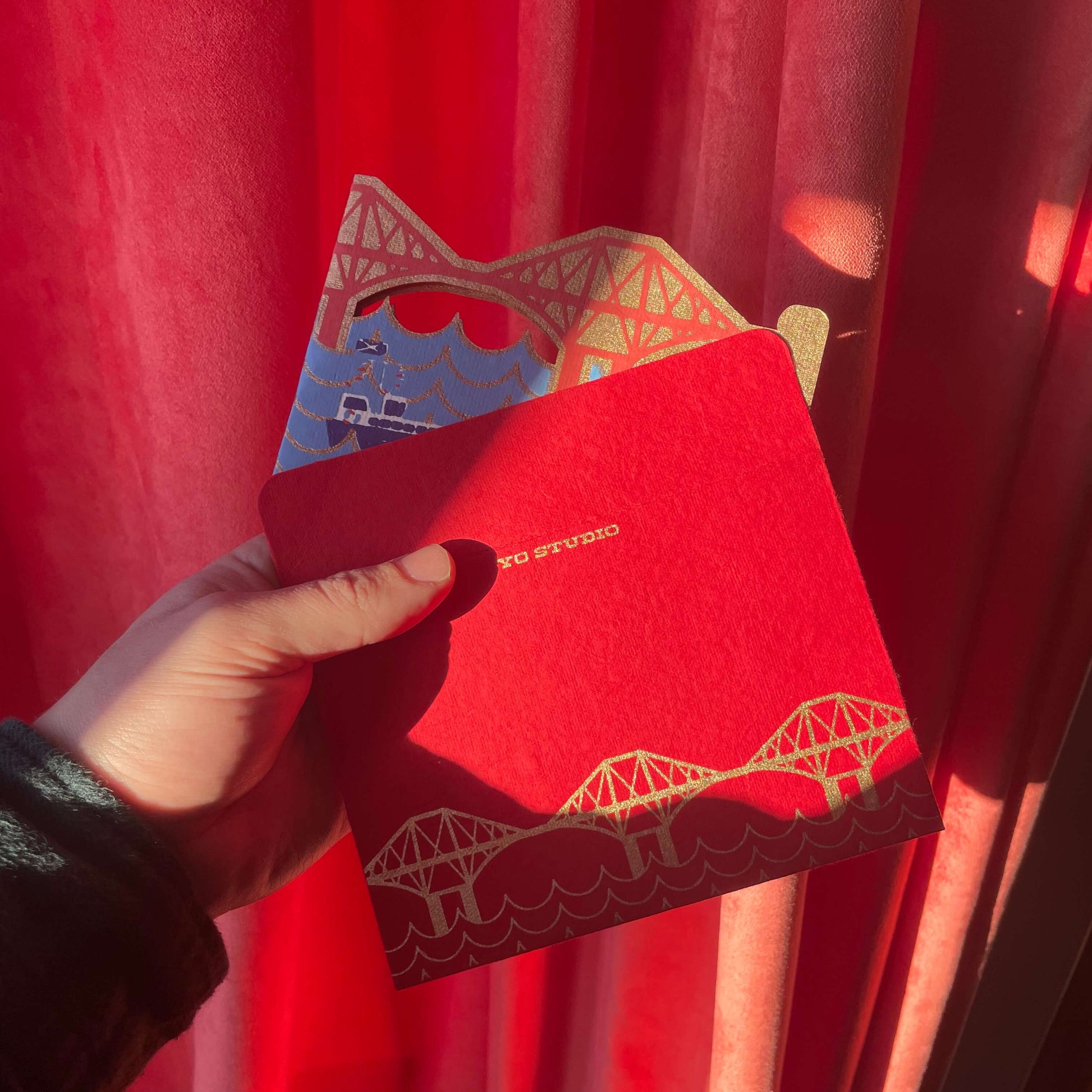 Red envelope with Forth Bridge design held by a hand against a red curtain