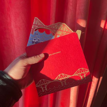 Red envelope with Forth Bridge design held by a hand against a red curtain
