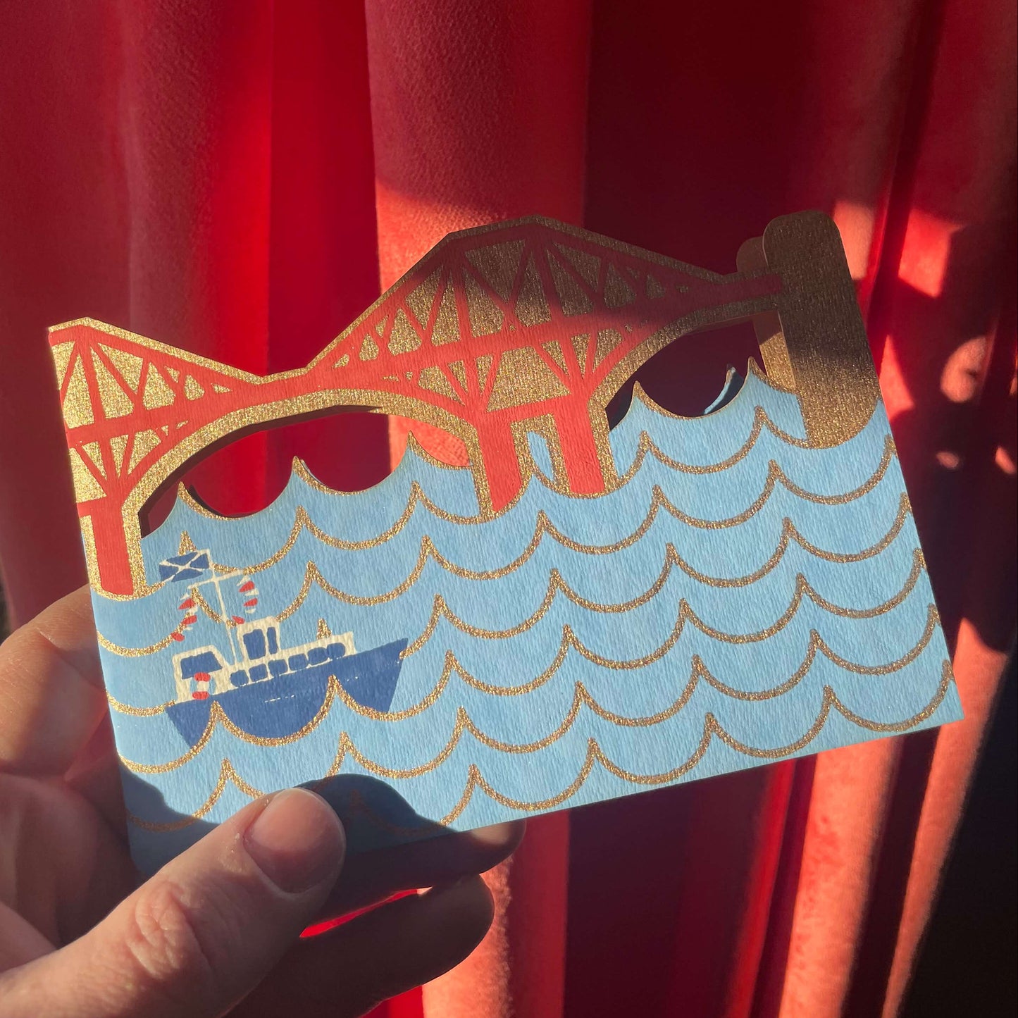 Card with a design of a Forth Bridge card  and waves held against a red curtain.