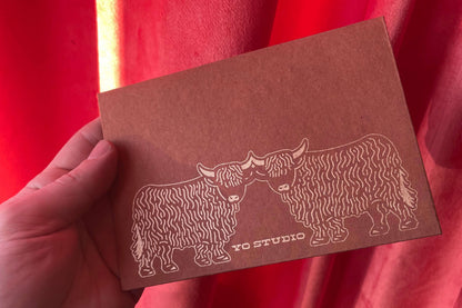 Hand holding a brown envelope with highland cow illustrations against a red curtain background