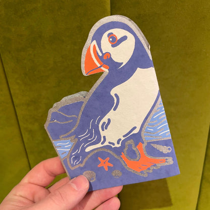Hand holding a decorative card with a puffin design against a green background