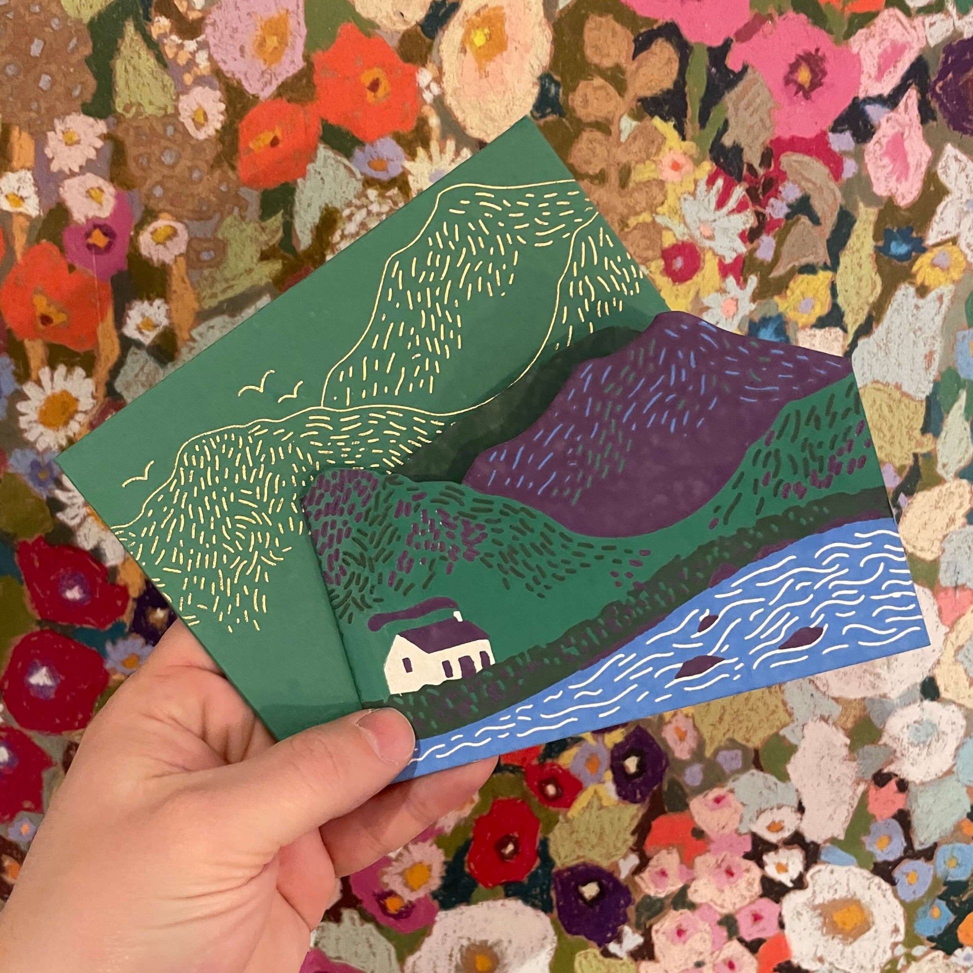 Hand holding a colorful landscape illustration against a floral background
