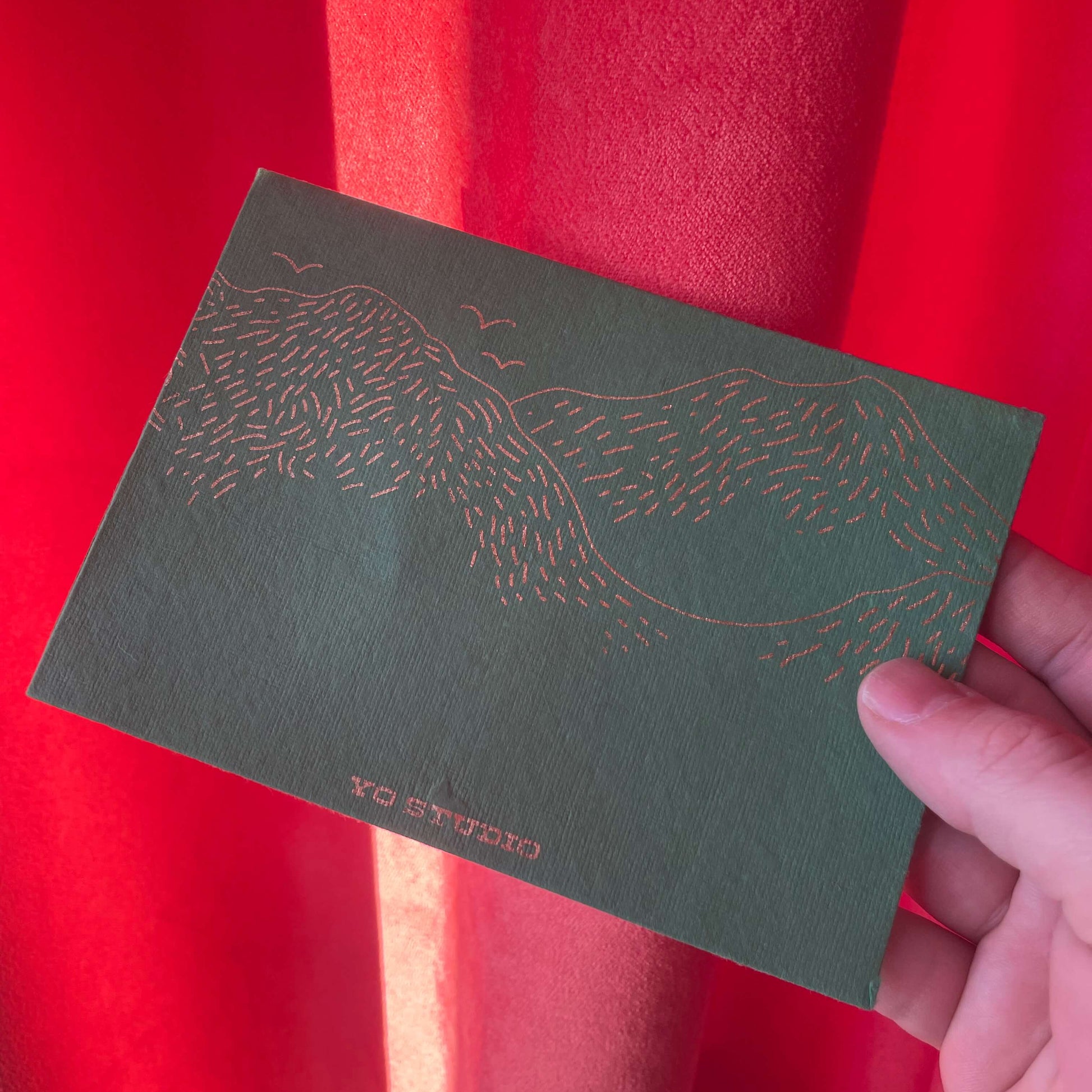 Green card with a landscape hill illustration held against a red fabric background